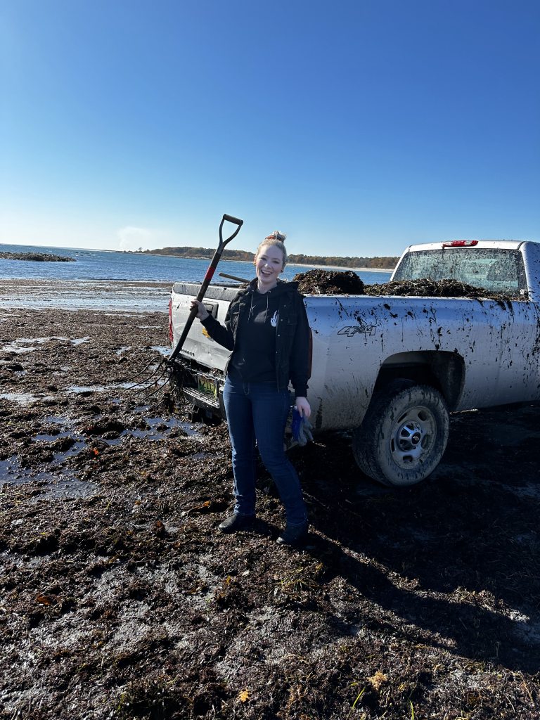 Woman smiling by a muddy pickup truck with a shovel, near a beach.