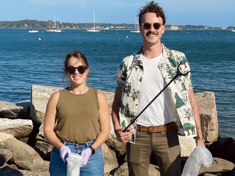 A man and woman holding trash bags on a rocky beach, ocean background.