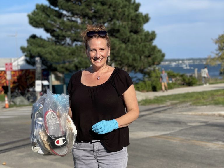 "Cheerful woman holding a bag of trash, standing by the seaside on a sunny day."