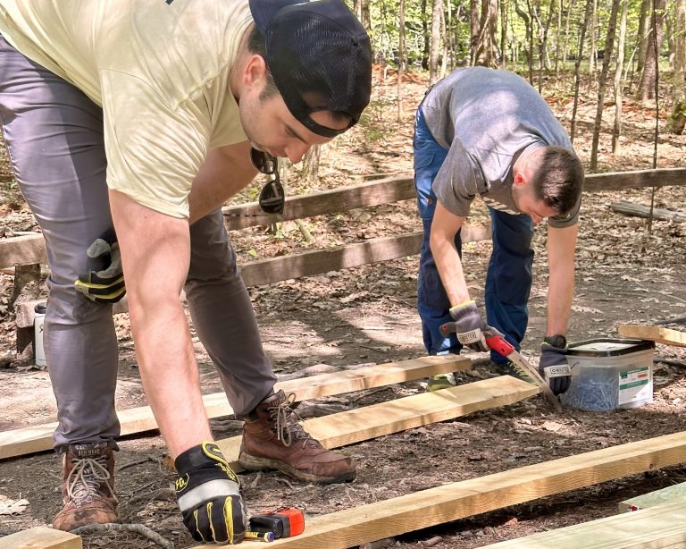 Two people measuring and cutting wood outdoors.